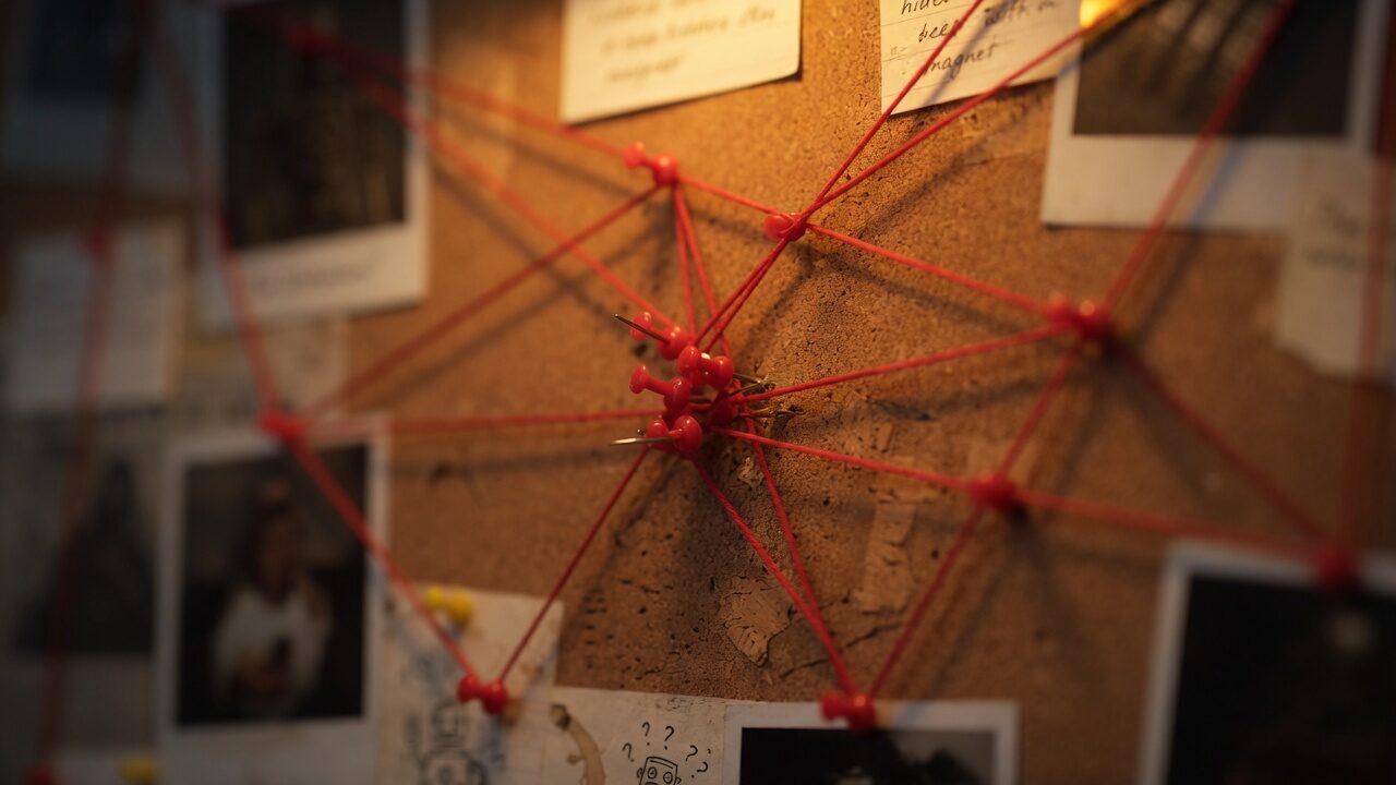 Close-up of red pushpins and taut crimson string radiating across a corkboard, with Polaroids and scribbled notes at the edges.